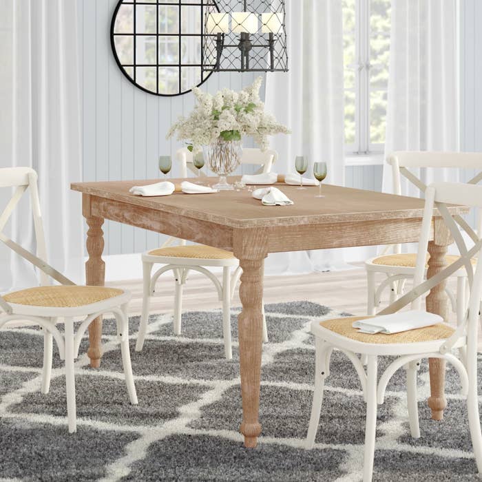 Brown wood farmhouse-style kitchen table with four white chairs around it and a vase of flowers on top