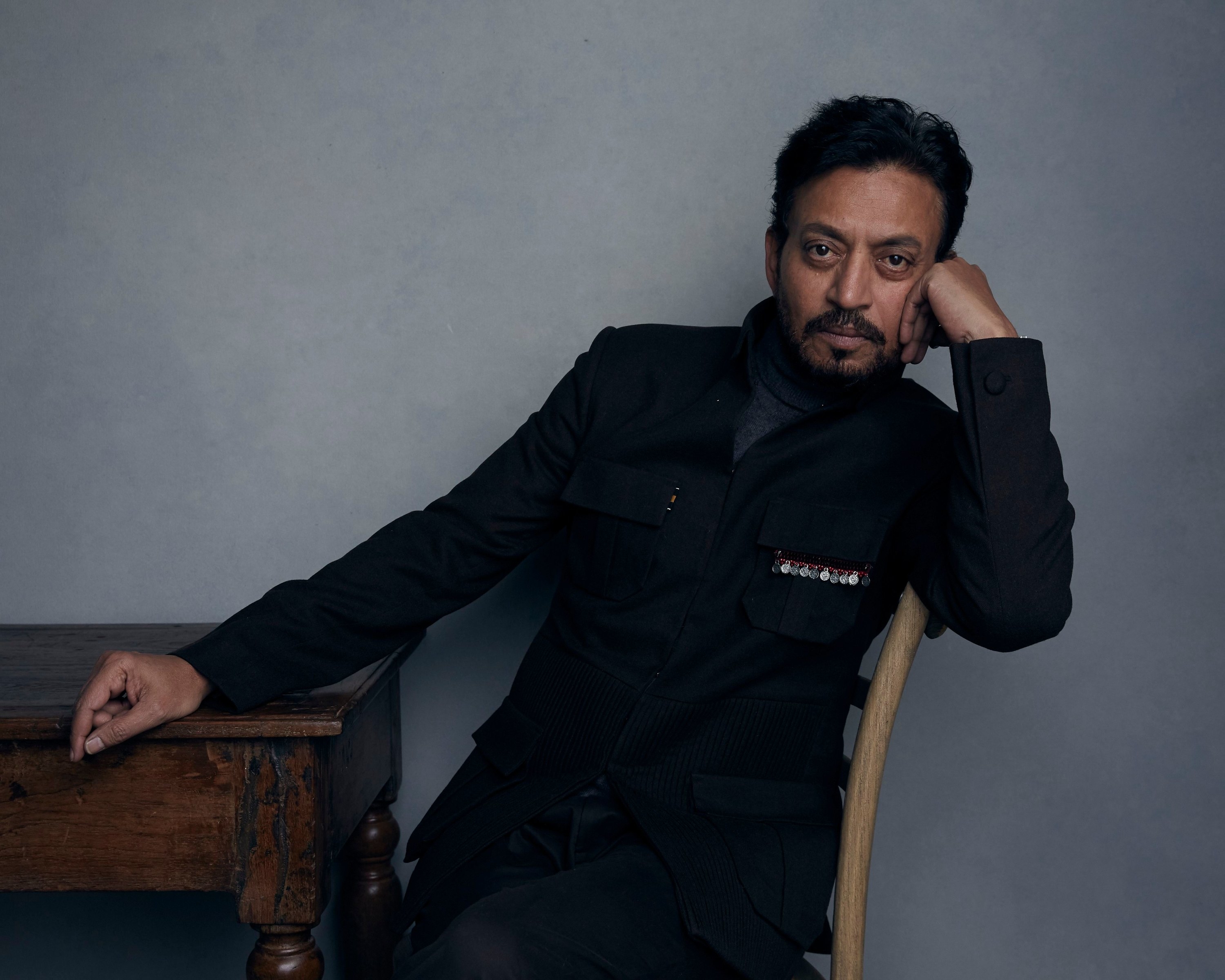 Khan in a black suit seated at a table, staring at the camera