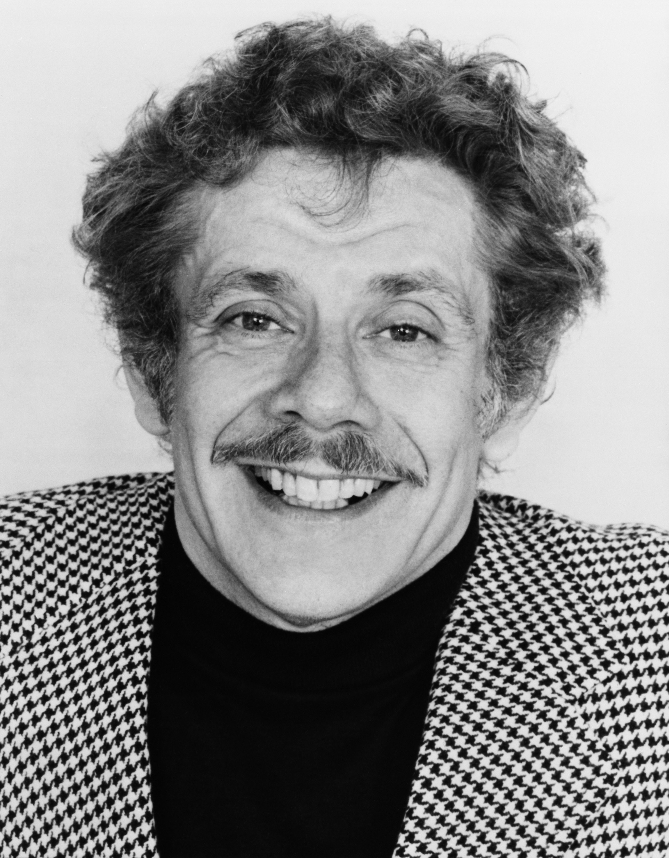 Man with curly hair and a checked suit smiling at camera