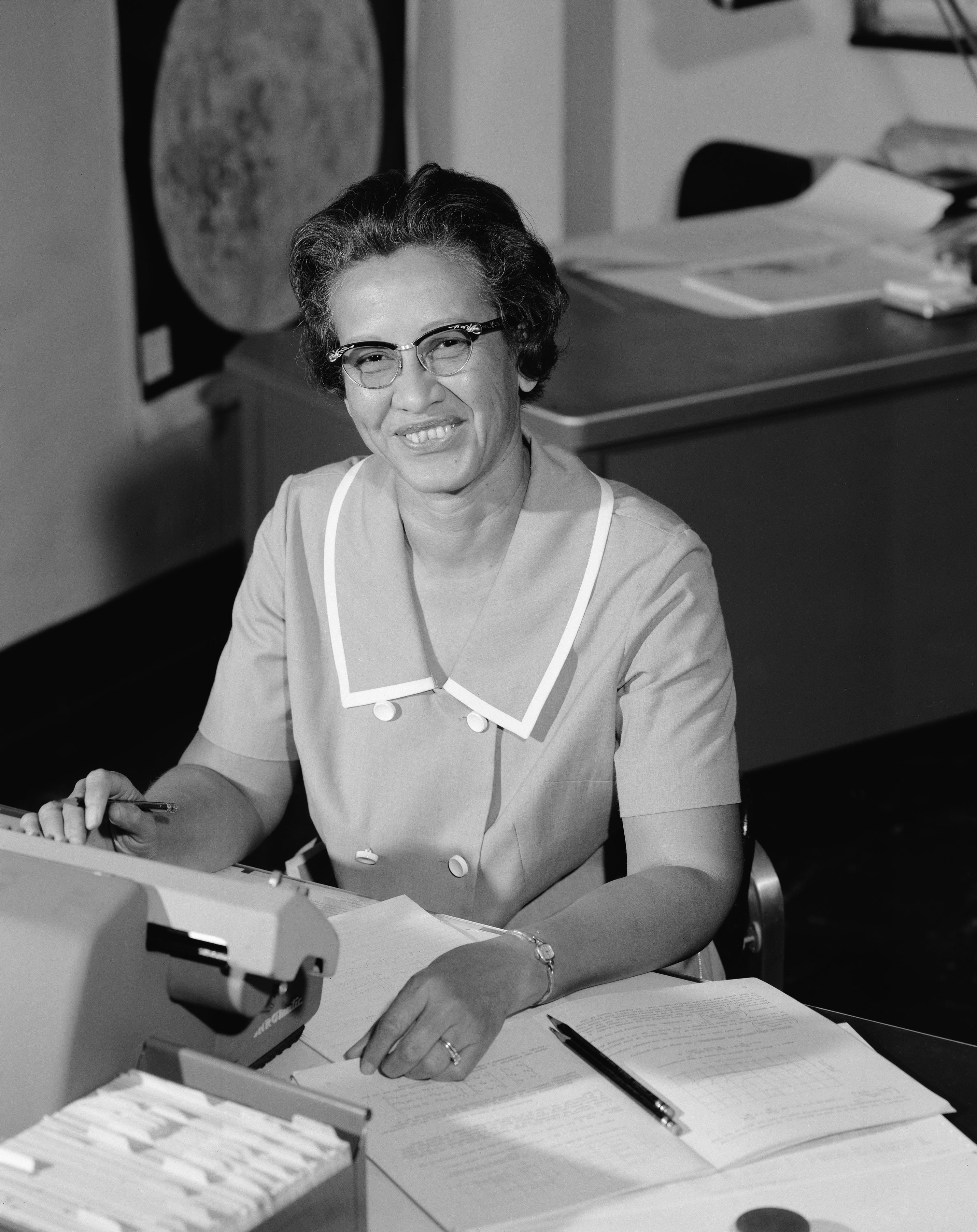 A woman in glasses and a short sleeve suit at a desk with paper, typewriter