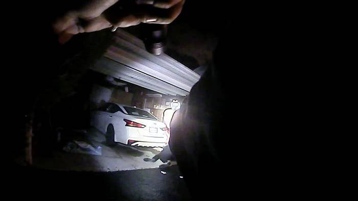 A still from body camera footage shows a police officer pointing a gun and flashlight at a body, which lies on the ground in front of him outside a garage