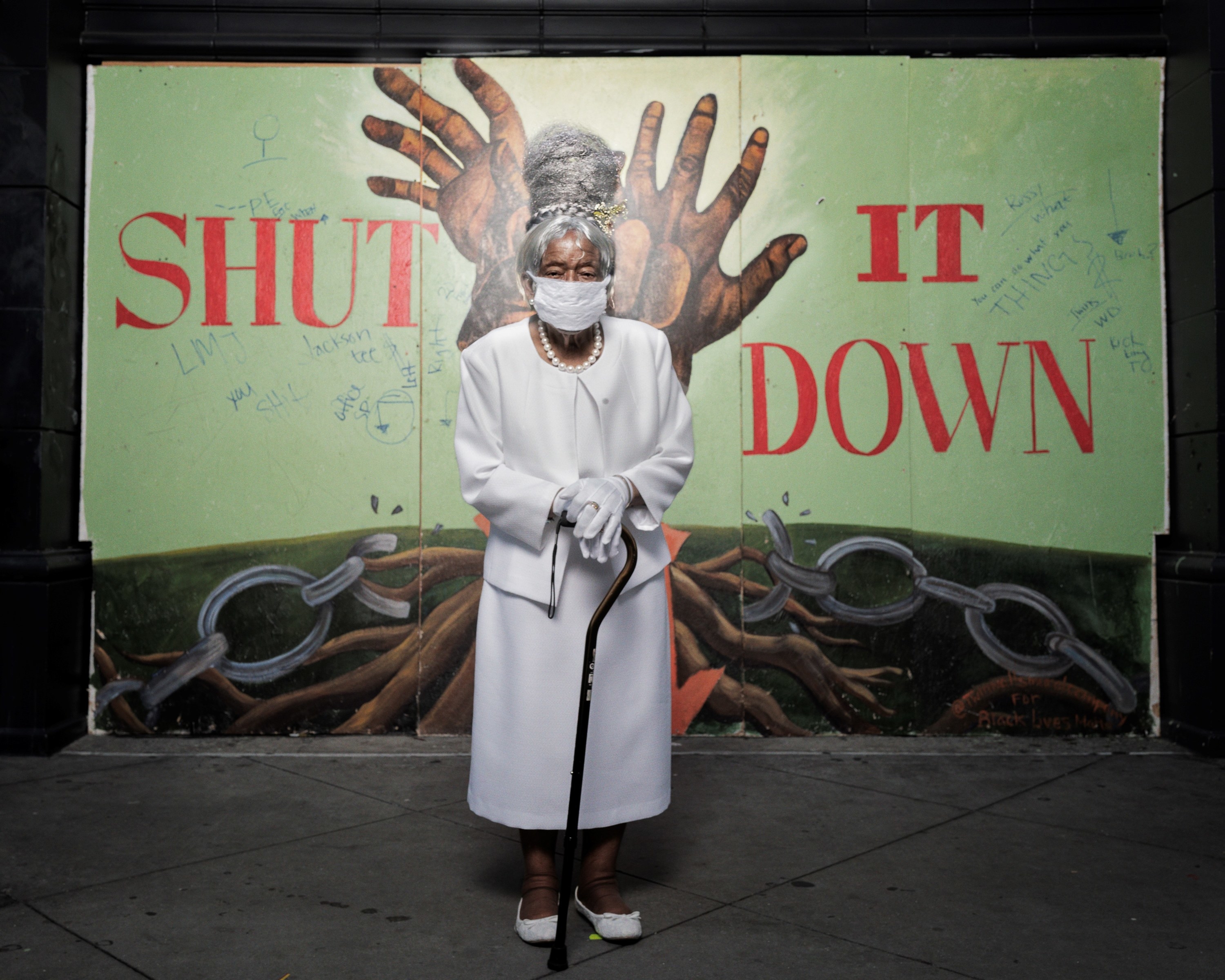 An older woman with a cane and long coat in front of a mural of hands breaking free from chains
