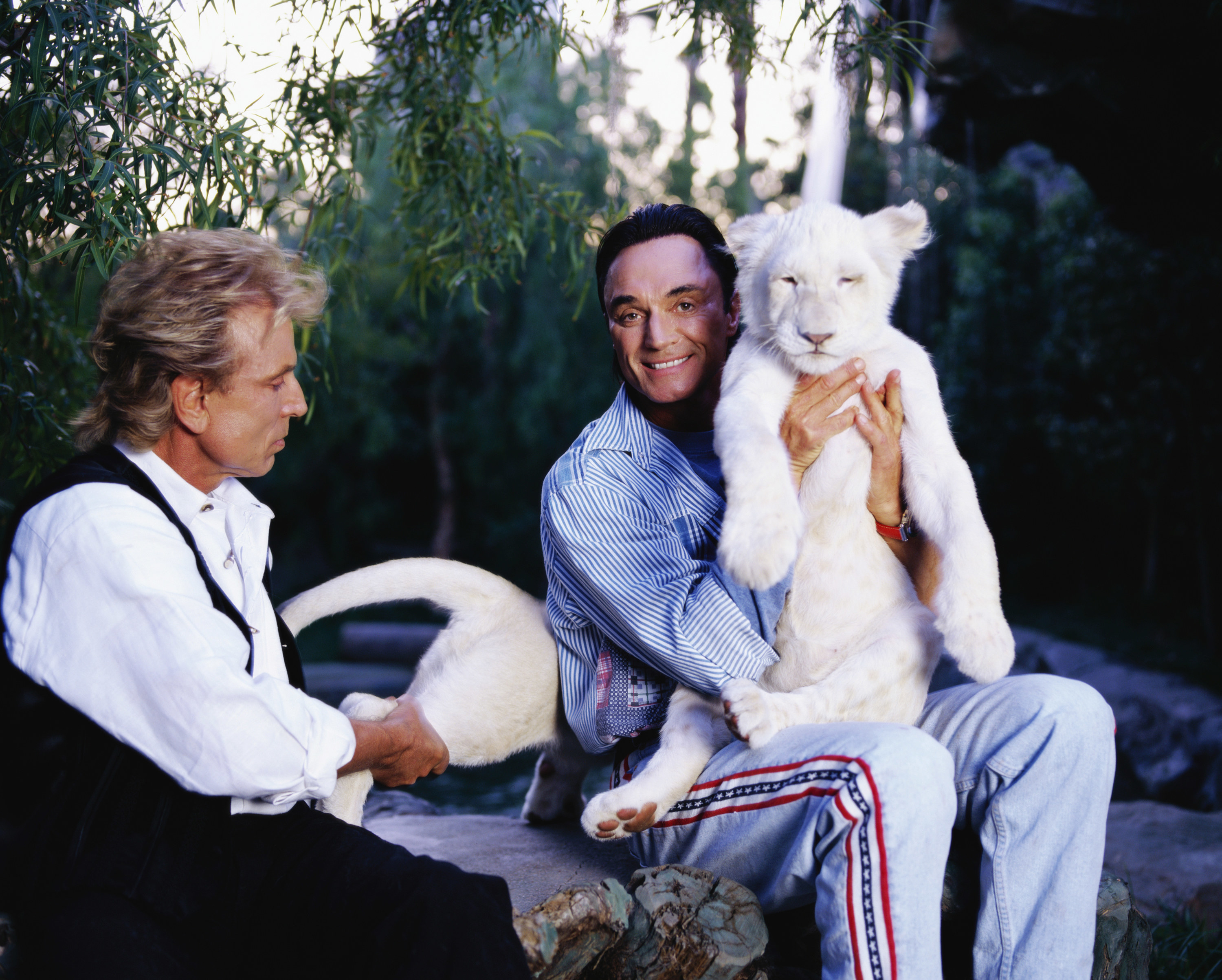 Two men seated outside, one man has a small white tiger on his lap that he is holding up for the camera