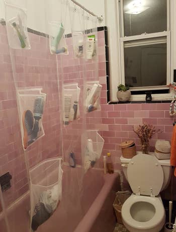 Reviewer uses clear shower curtain with pockets to store brushes and grooming products in their pink bathroom