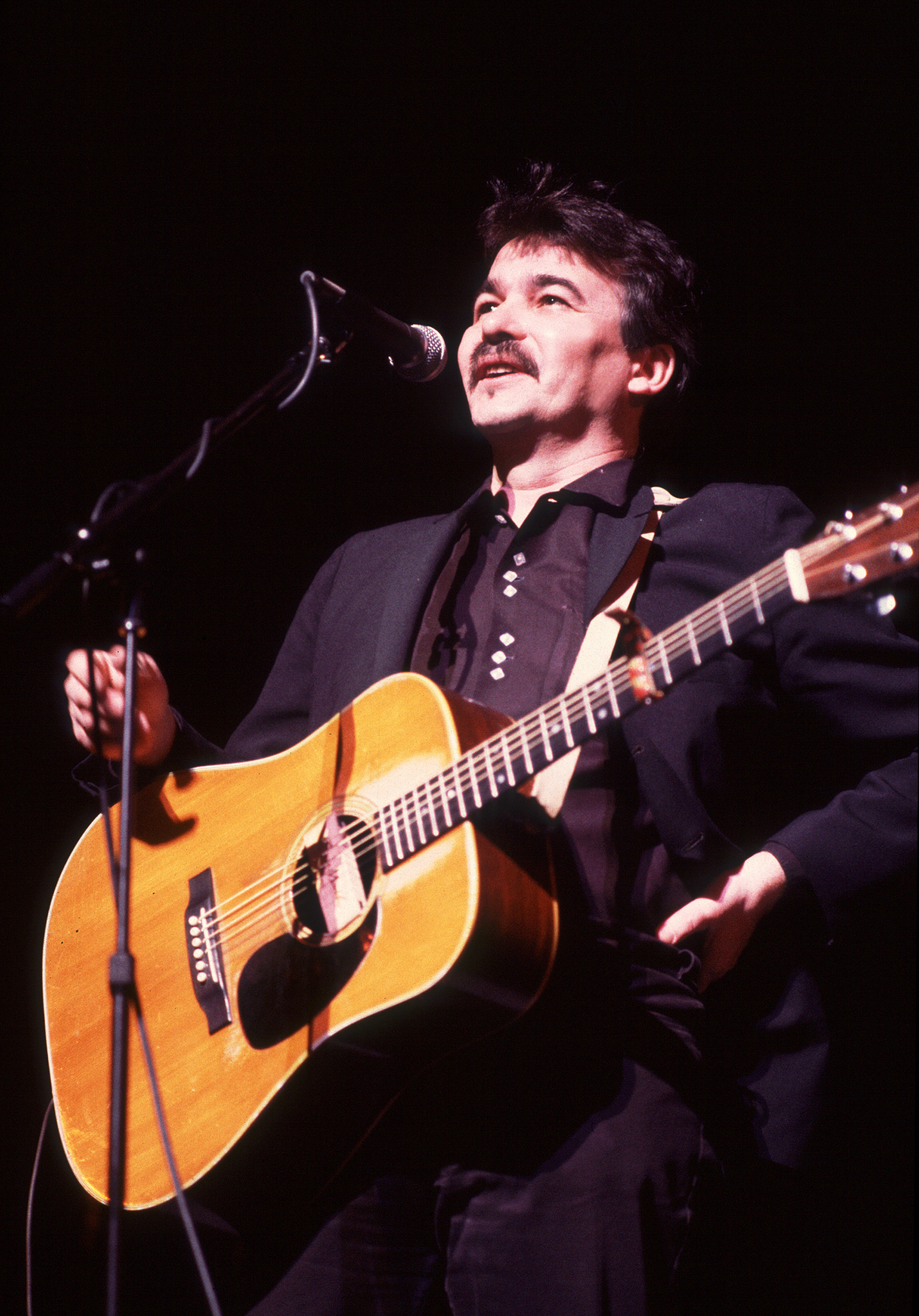 Prine on stage with bright lights on him holding a guitar