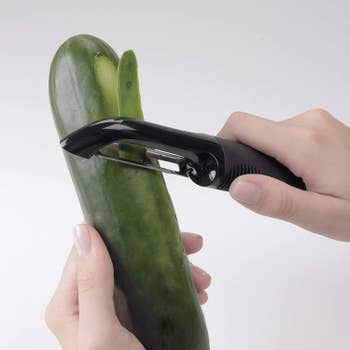 Hands holding the black peeler and a zucchini, peeling the side of it