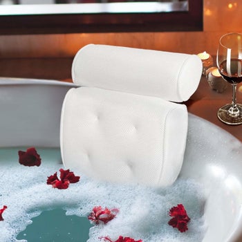 Plush white bath pillow on back of white tub with bubbles and rose petals inside