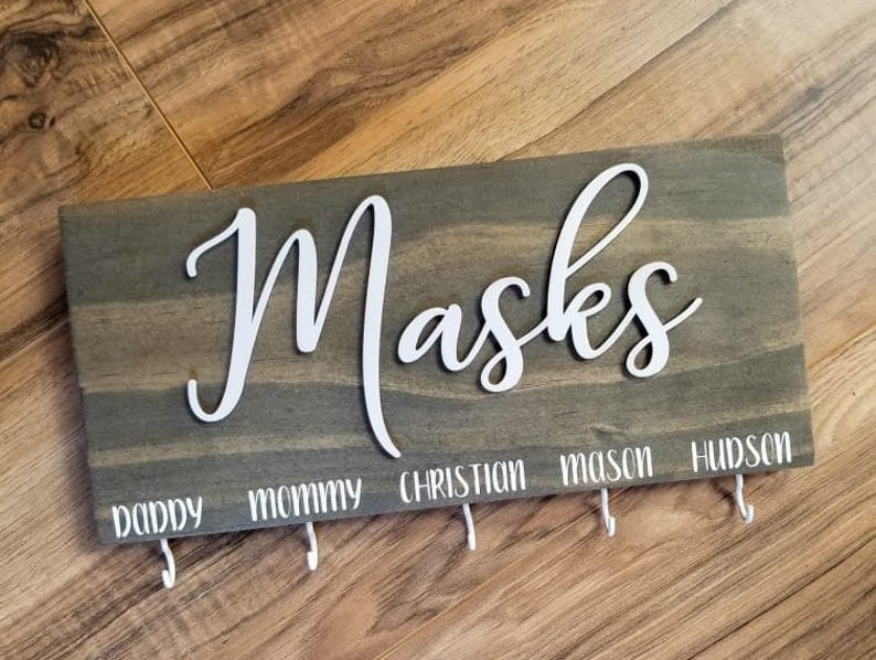 A wooden sign that says &quot;Masks&quot; in white script with five small hooks and names above them 