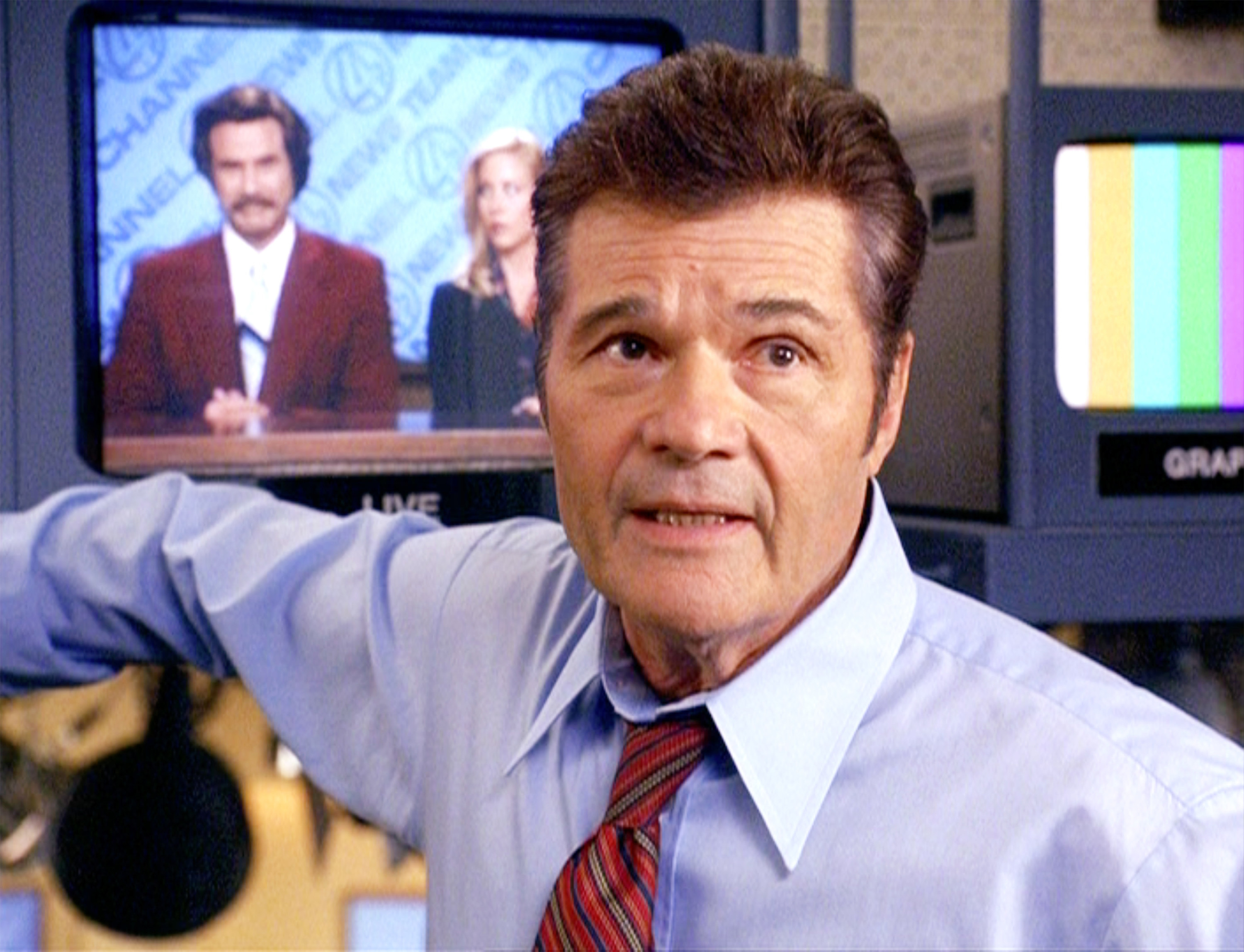 Fred Willard in a shirt and tie in front of televisions on the Ron Burgundy set