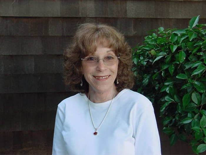 An older woman smiles at the camera and stands in front of a plant outside