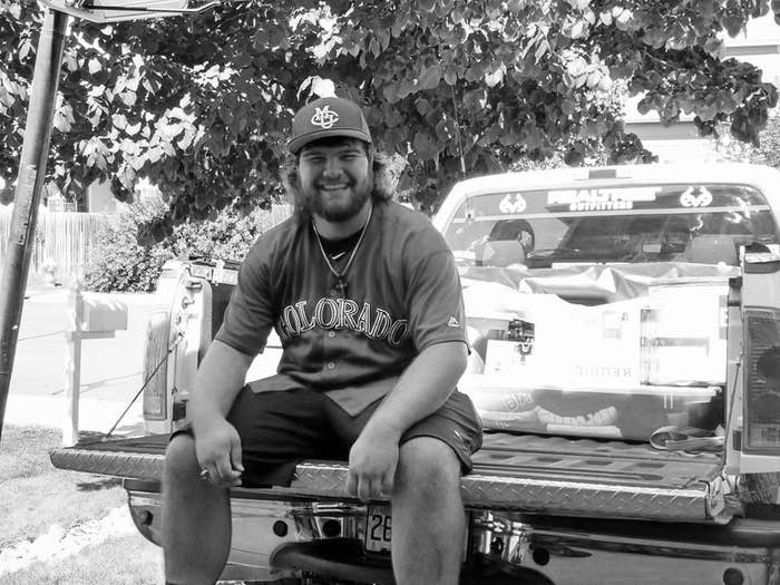A young man sits in the bed of a pickup truck, wearing a baseball jersey and CMU cap