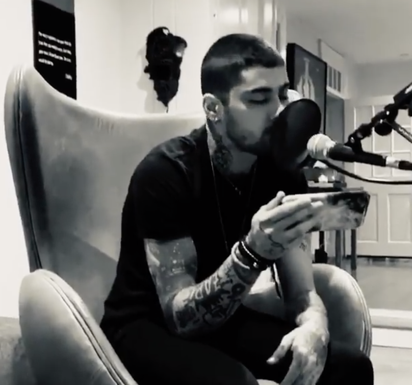 a man with a lot of tattoos sits on a chair, singing into a microphone, looking at his phone