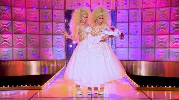 Drag queens Pearl and Trixie Mattel wearing a conjoined white and gold gown, dressed as child beauty pageant contestants