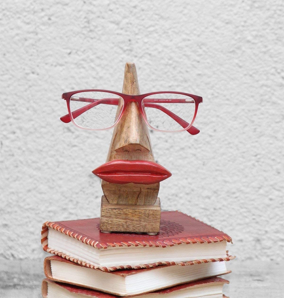A woden nose shaped eyeglasses holder with red lips resting on a pile of red books 