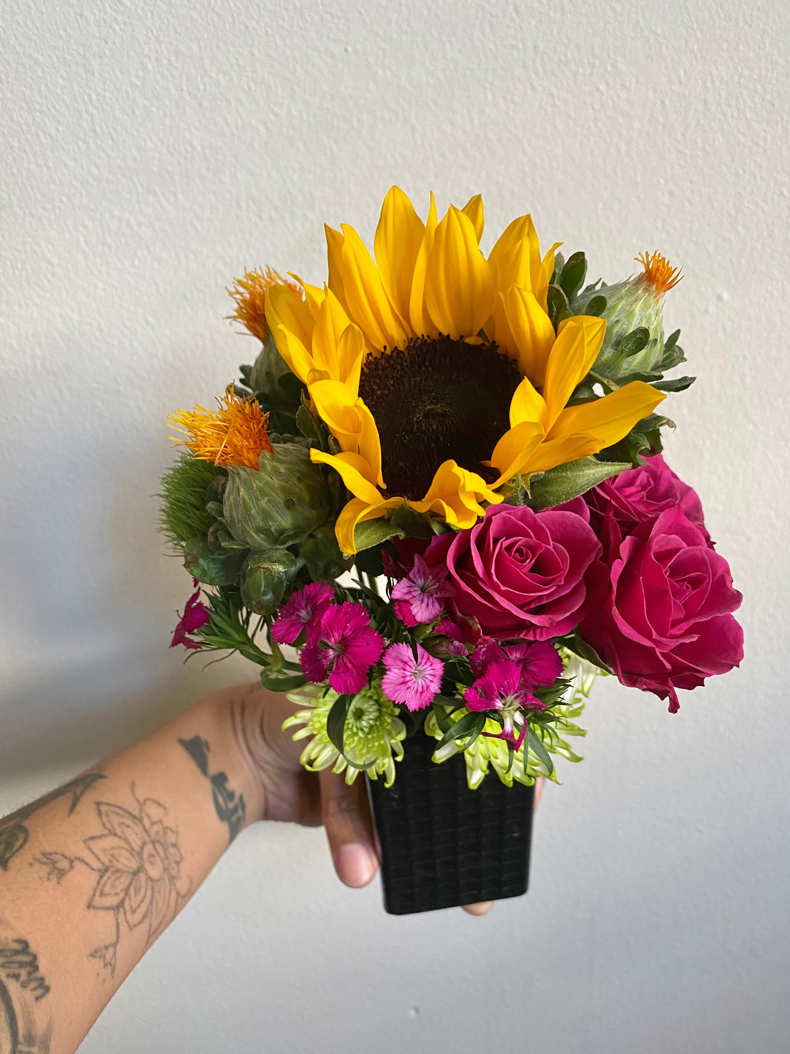 model&#x27;s hand holding an arrangement with pink roses, sunflowers, and more