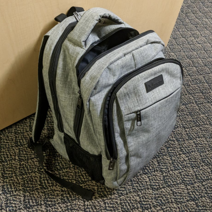 Reviewer image of gray backpack upright on floor showing multiple front pockets, laptop sleeve, and storage 