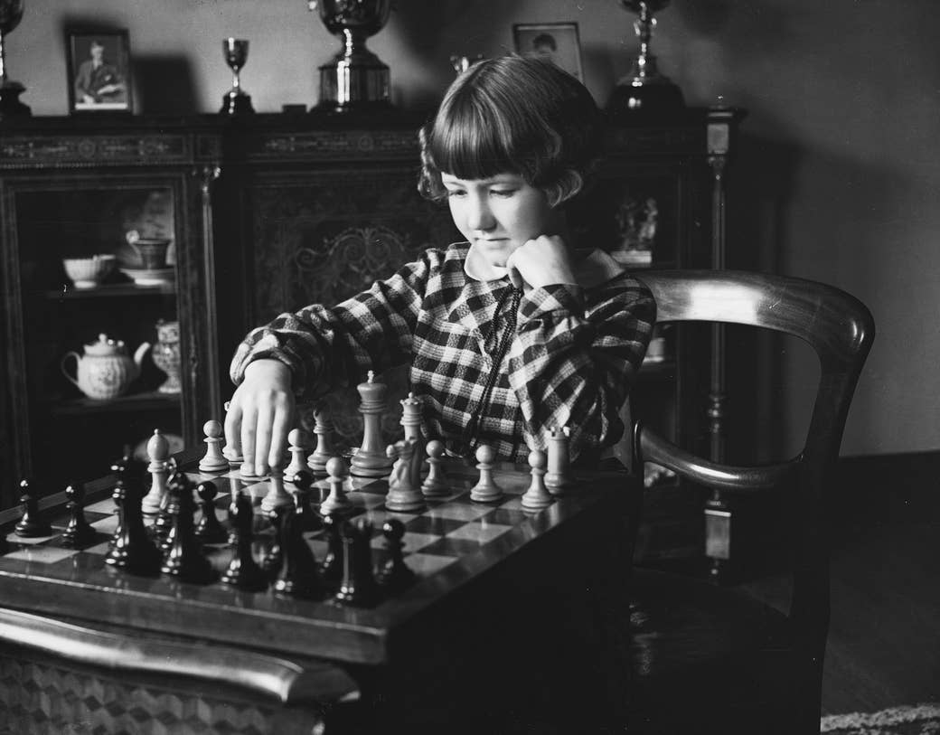 A young girl in a checkered dress plays chess in front of trophies on a shelf