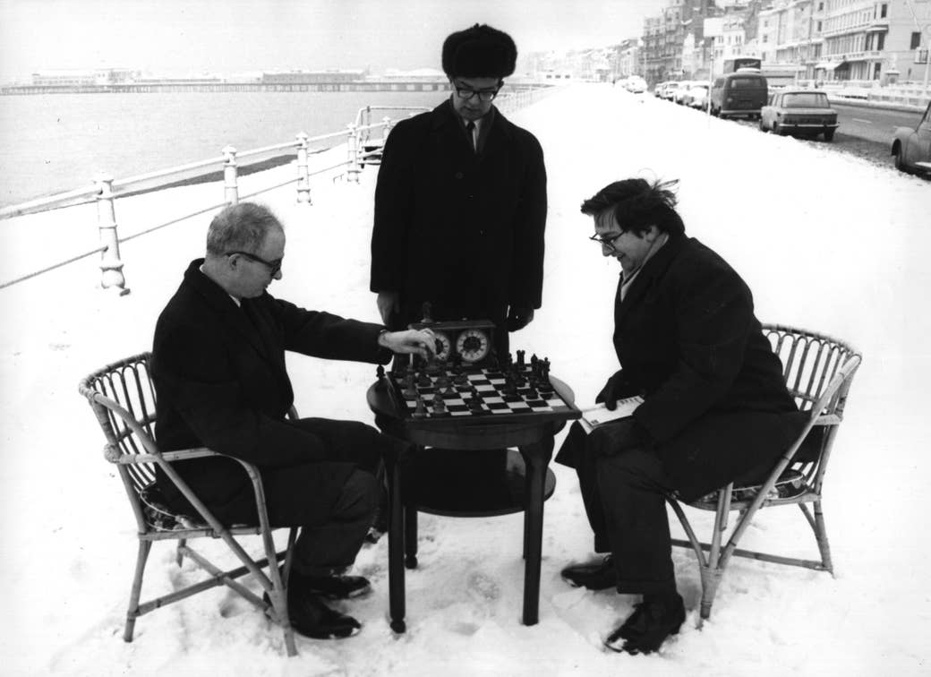 Two men seated in the snow play chess as one man, standing and wearing an ushanka, watches them play