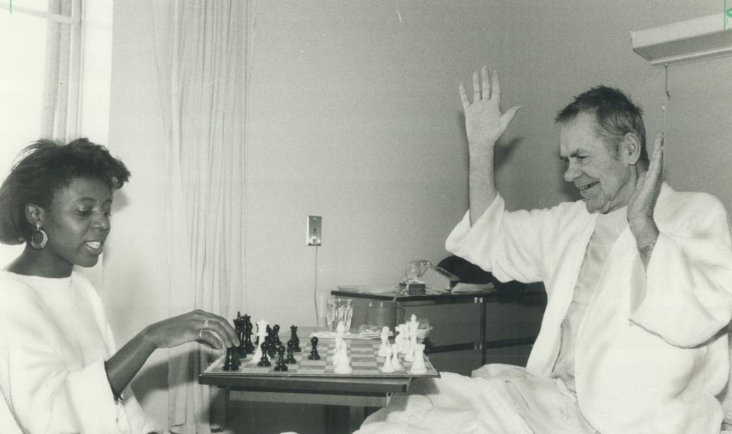 A young woman and an old man in a hospital bed playing chess.