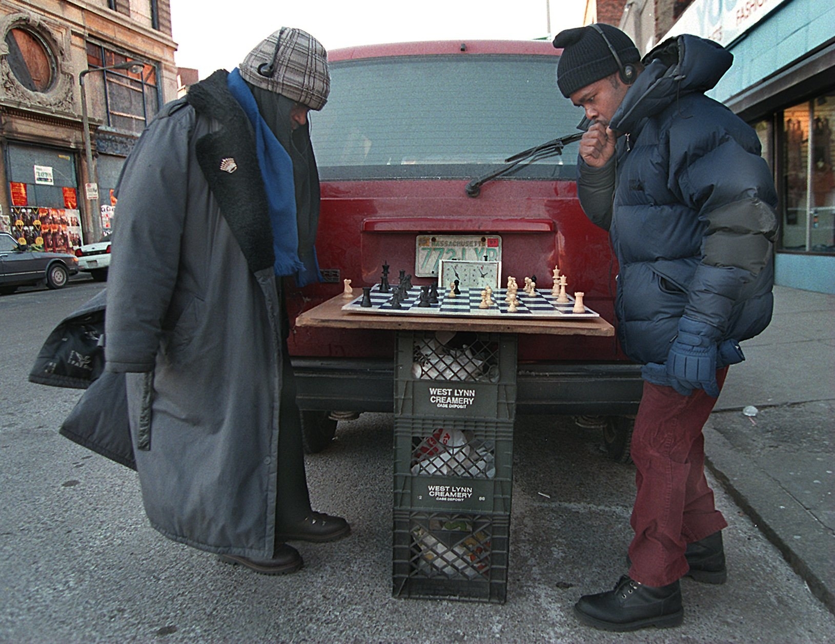 Two Black men in overcoats and hats play chess on three stacked-up milk crates behind a maroon minivan