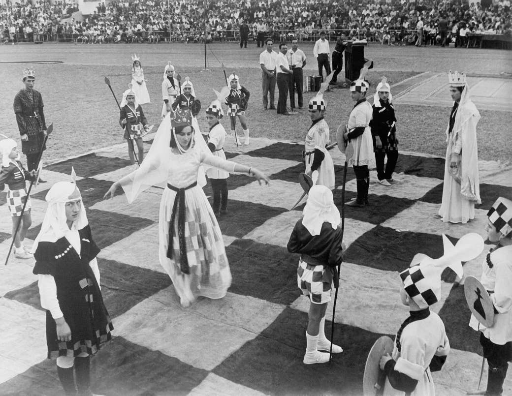 A woman dressed as a chess queen, with a crown and checkerboard dress, has her arms outstretched, standing on a giant chessboard and surrounded by other people dressed as pieces