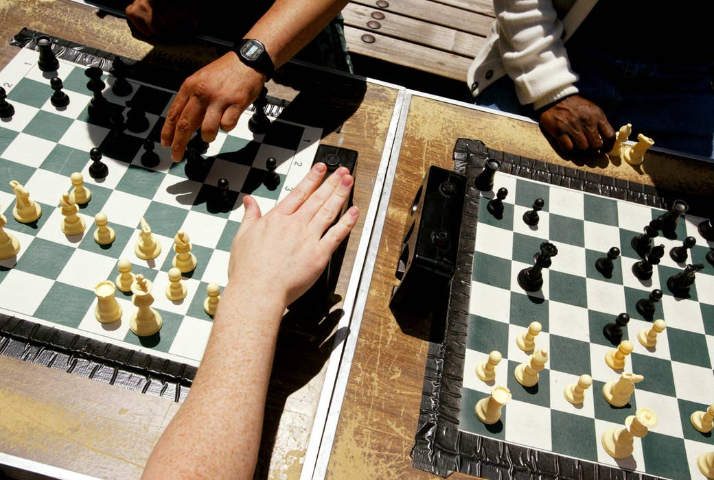 A hand depresses a chess clock on a table between two chessboards in the sunlight. 