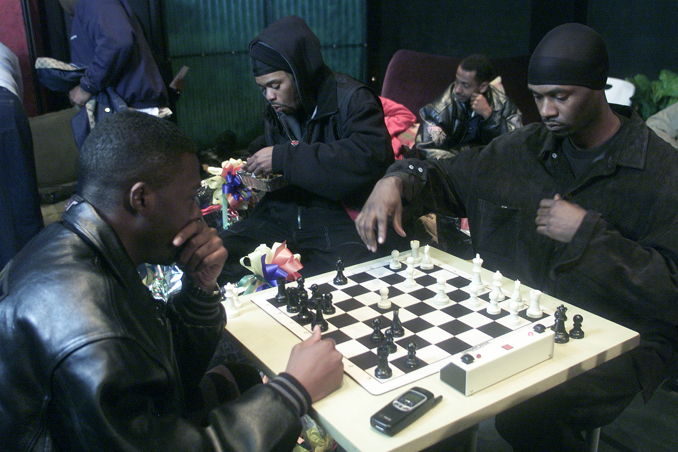 Four men hang out in a backstage green room; two of them in the foreground play chess