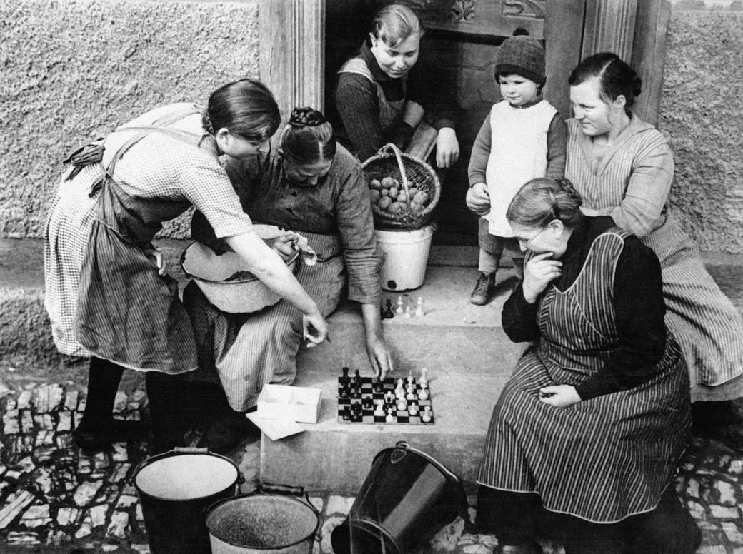 Five women and a toddler, all in aprons and old-fashioned clothing, huddle around a chessboard on a stoop