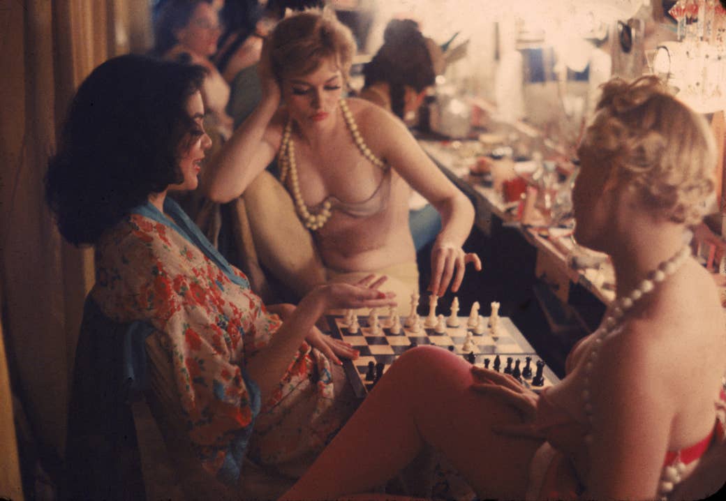 Three women in a backstage green room, wearing robes, underwear, and pearls, sit around a chessboard