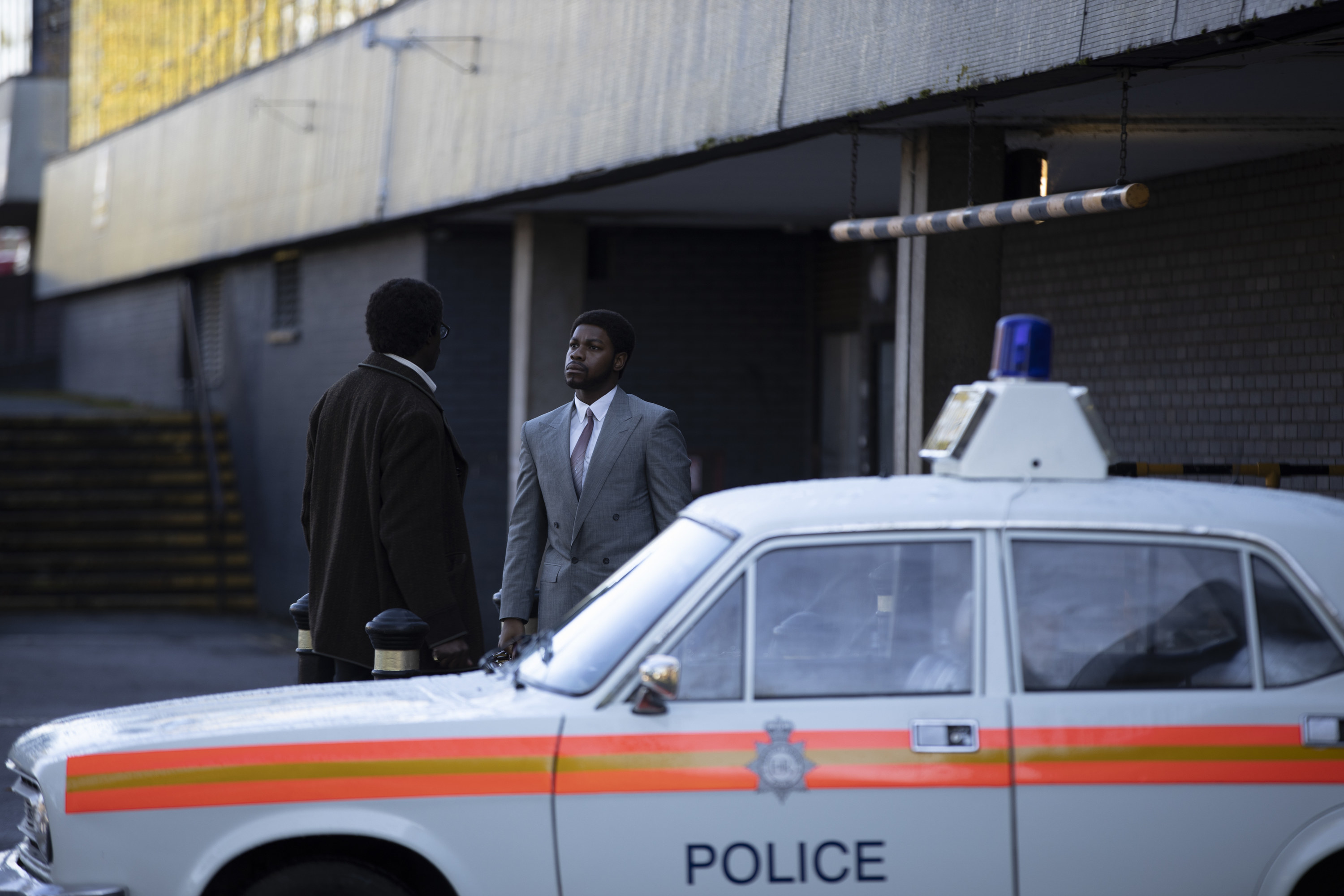 John Boyega in &quot;Red, White and Blue&quot; dressed in a suit, standing in front of a police car