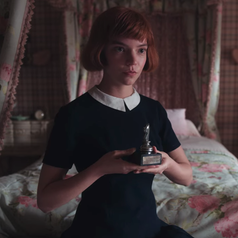 Beth sitting on her bed while holding a chess trophy; she is wearing a short-sleeved navy blue dress with a Peter Pan collar