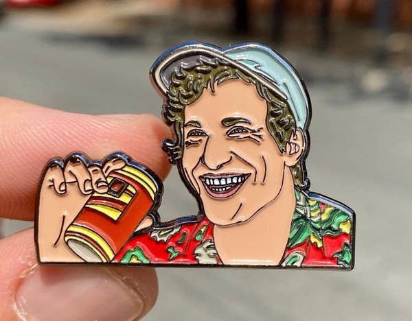 A label pin of Andy Samberg in his costume from the movie Palm Springs, smiling and holding a beer can