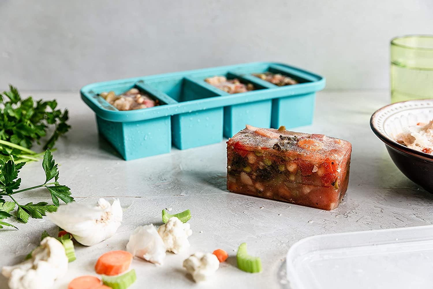 The five-compartment teal container that looks like a large ice cube tray, with a perfectly rectangular block of frozen soup next to it 