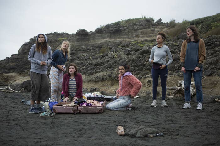 Six teen girls stand and kneel on a beach on an overcast day, a suitcase of clothes open before them