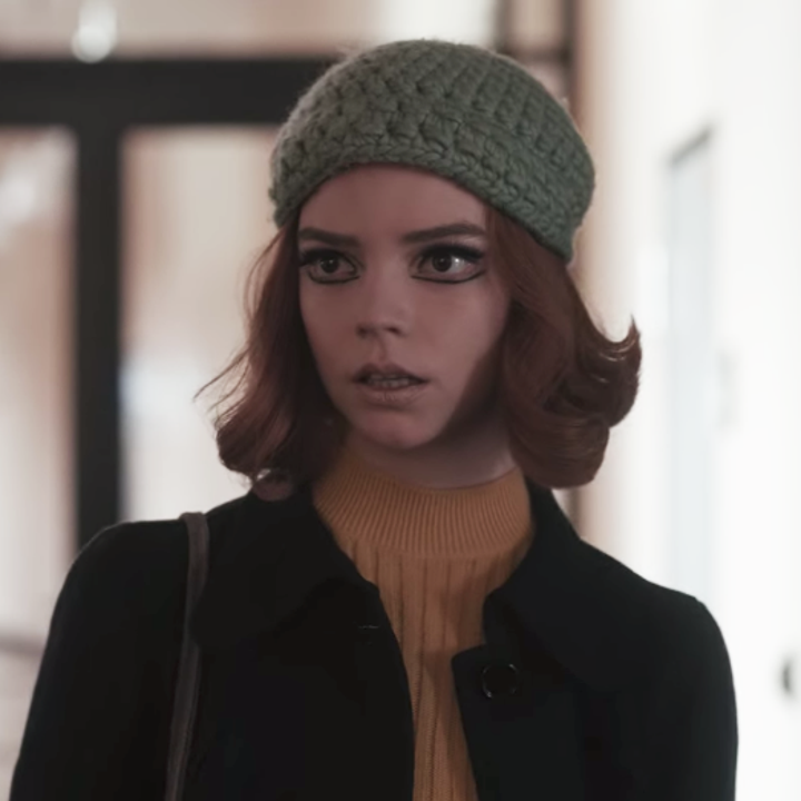 Beth standing a hallway at a school; she is wearing a grey beanie with a mustard yellow turtleneck and a black coat; her makeup features a nude lip and bold eyeliner