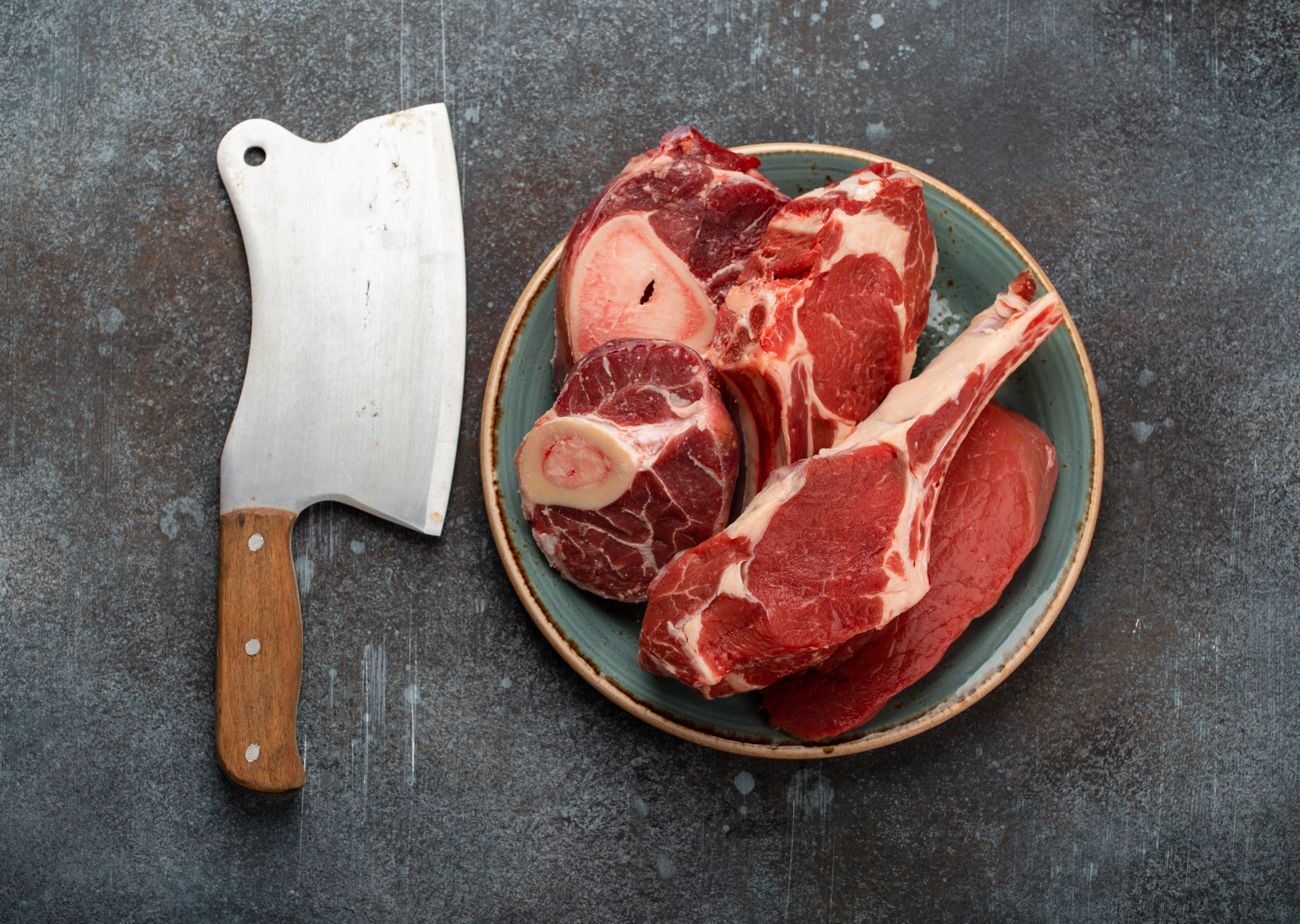 Raw cuts of meat on a plate next to a butcher&#x27;s knife