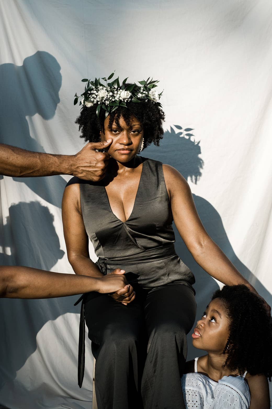 A Black woman in a flower crown with a young girl by her right side and two male hands reaching out to her from the left. 