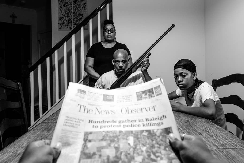 A newspaper in the foreground with a headline about police killings, with a Black family at their table in the background. The man is holding a rifle. 