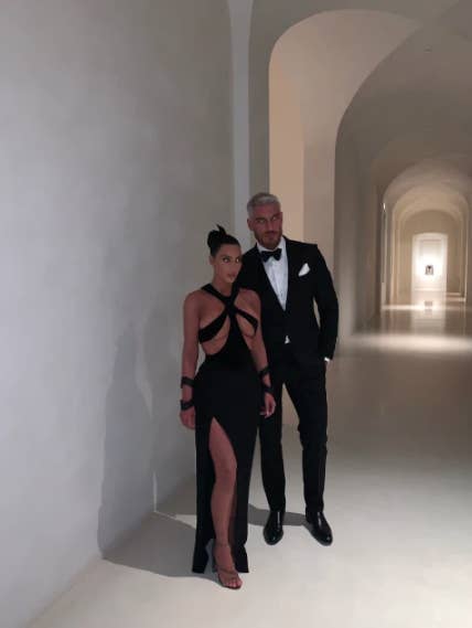Kim, who's wearing a dress with several cutouts, poses for a photo with another person, who's wearing a tux, in Kim's very minimalist hallway
