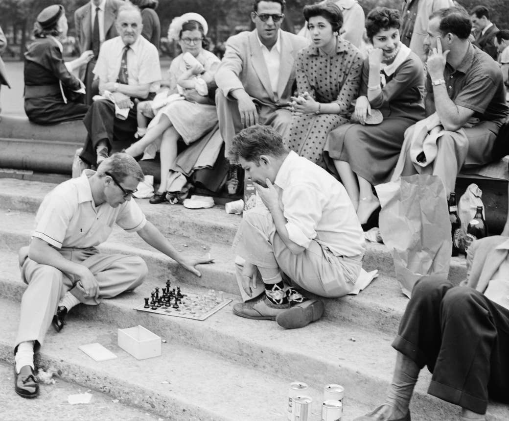 A group of people sitting on stairs at a park surround two men kneeling and playing chess
