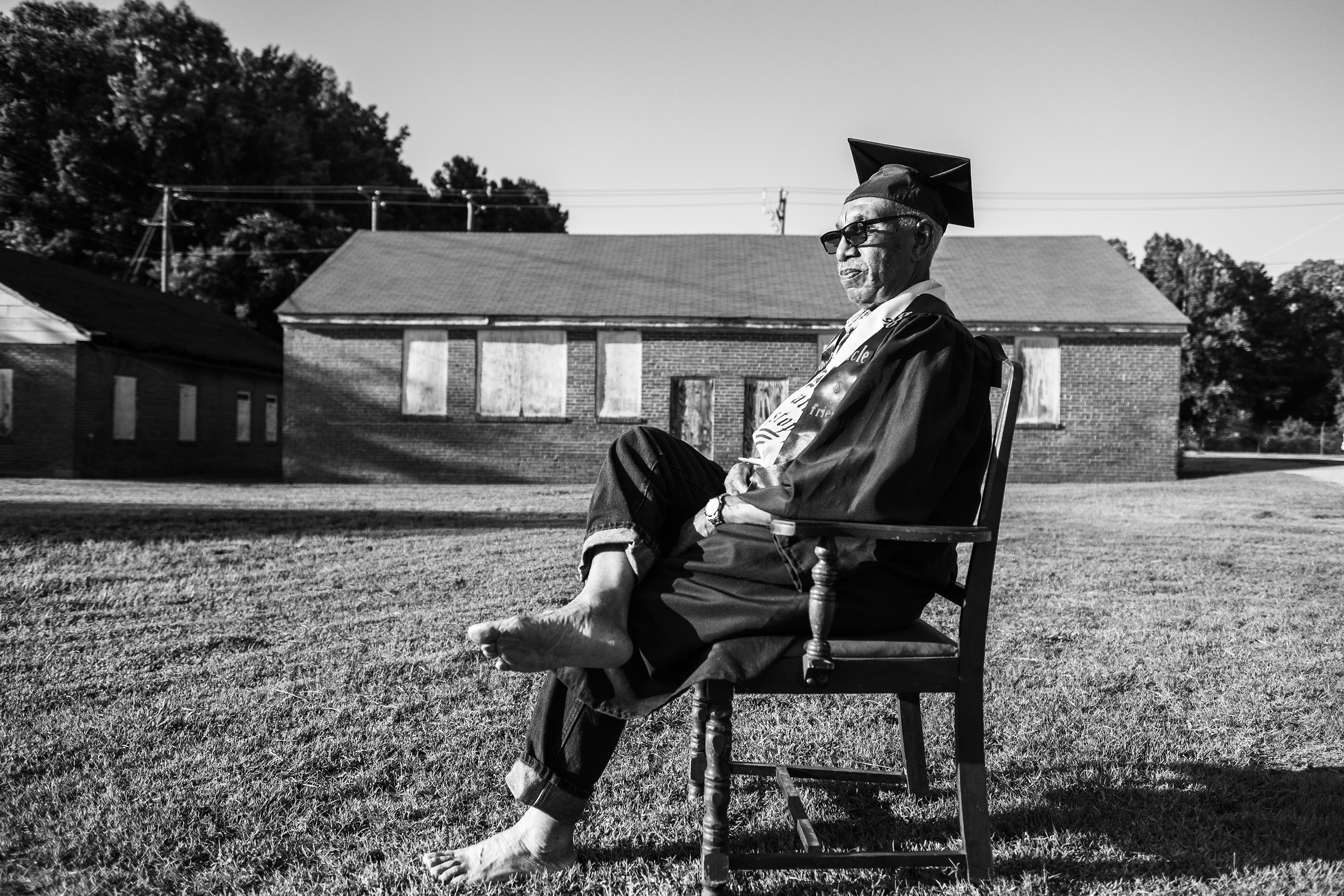 A Black man in graduation robes sits barefoot on a chair in a backyard