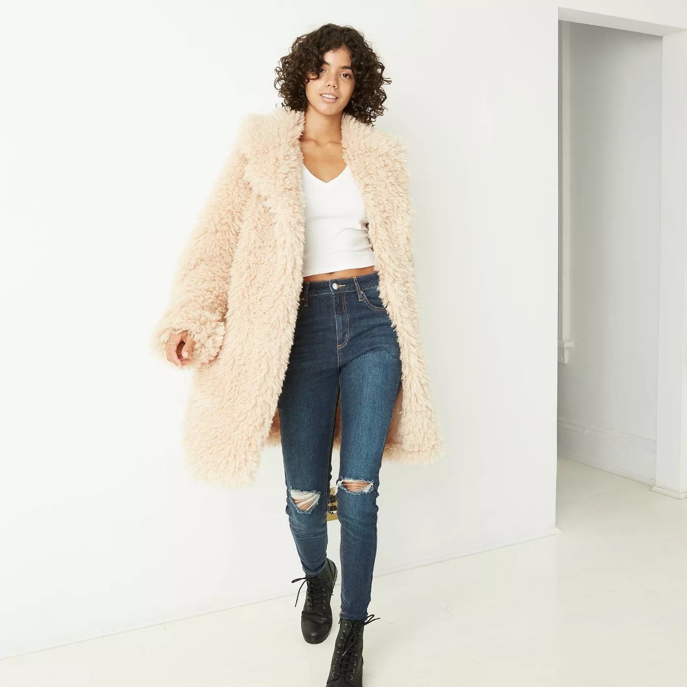 Light beige jacket that is knee length. Model is wearing jeans, white tank, and black combat boots. 