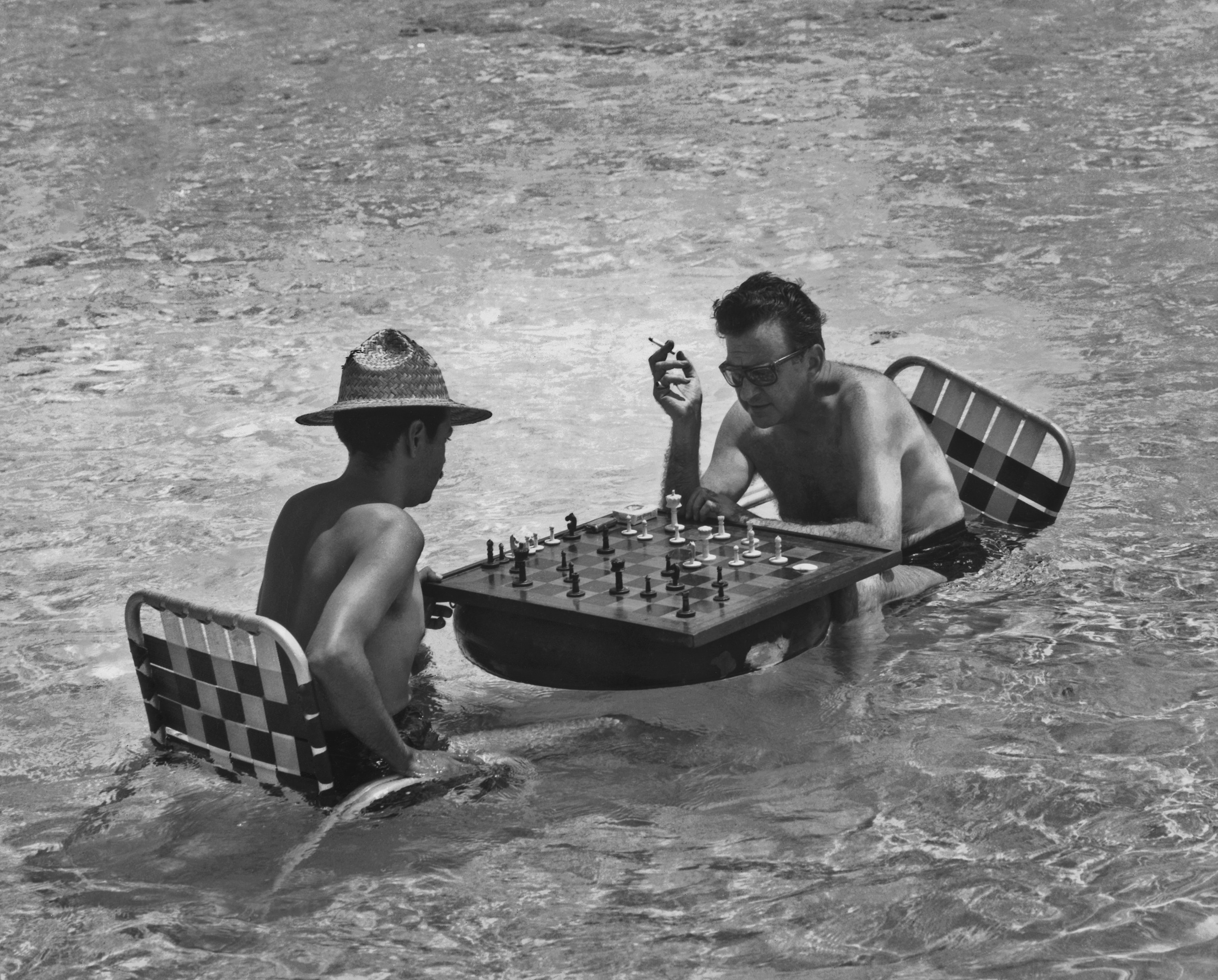 Two men sitting in the water in beach chairs play chess on a board that floats on an inner tube