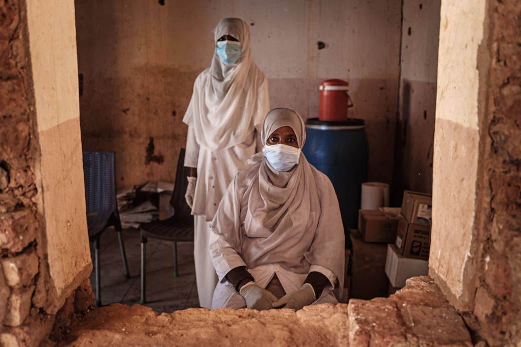 Two women in veils and protective equipment in a small room, seen through a window without glass