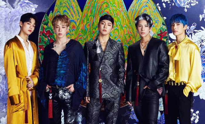ACE poses together wearing a mixture of suits, robes, and silk shirts with a colorful background behind them