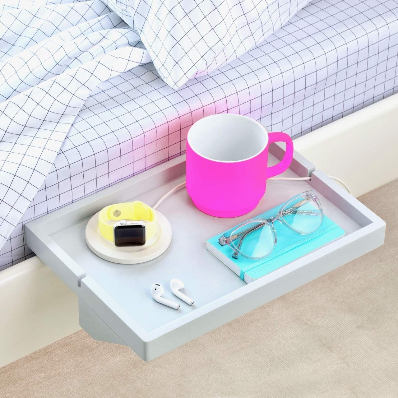 the shelf attached to the side of a bed with a mug, air pods, glasses, and other items on it 