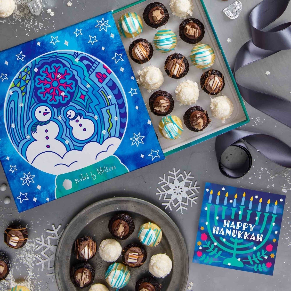 box with blue and pink snowman design with colorful mini cupcakes inside and a card with a menorah that says happy hanukkah