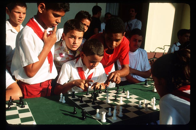 Boys in red and white uniforms huddled around a green and white chess board. 