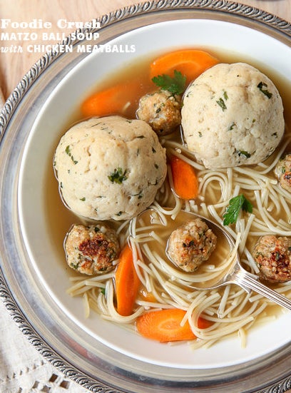 A bowl of soup with thin, long noodles, chicken meatballs, matzo balls, and carrots in chicken broth.