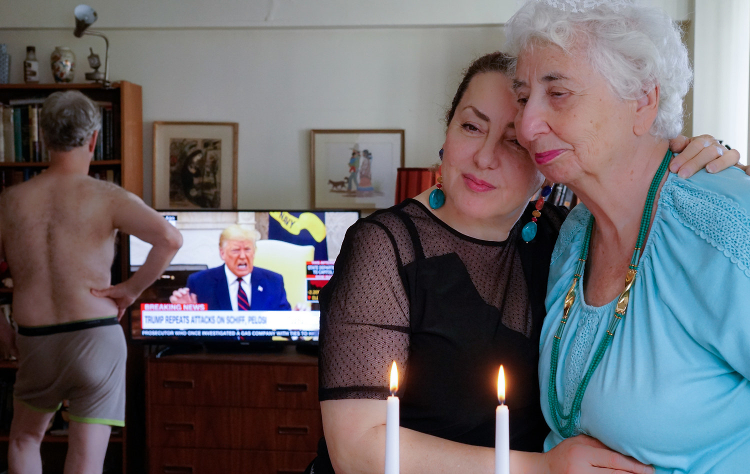 A man in his underwear watches Trump on TV while two women embrace in the foreground